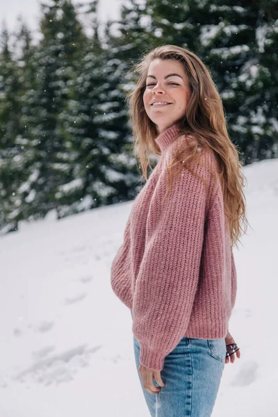 Egy fiatal, gyönyörű, hosszú hajú lány rózsaszín pulóverben pózol egy téli erdőben a hegyekben. — Stock Fotó