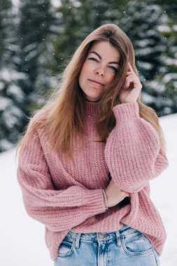 Genç, güzel, uzun saçlı pembe kazaklı bir kız dağlardaki bir kış ormanında poz veriyor.