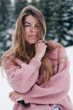 Genç, güzel, uzun saçlı pembe kazaklı bir kız dağlardaki bir kış ormanında poz veriyor.