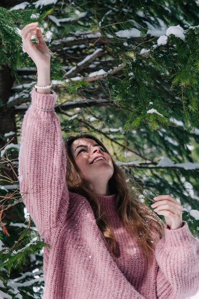 A young, cheerful girl sprinkled with snow under the branches of a spruce tree in the mountains — Stock Photo, Image