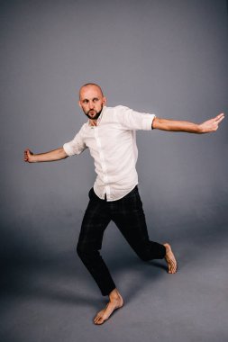 studio portrait of a bald guy with a beautiful beard barefoot in a white shirt and plaid pants dancing on a gray background