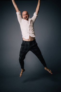 Beyaz gömlekli ve ekose pantolonlu, gri arka planda dans eden güzel sakallı, kel bir adamın stüdyo portresi.