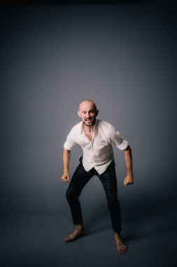 Beyaz gömlekli ve ekose pantolonlu, gri arka planda dans eden güzel sakallı, kel bir adamın stüdyo portresi.