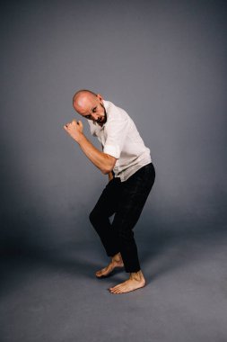 Beyaz gömlekli ve ekose pantolonlu, gri arka planda dans eden güzel sakallı, kel bir adamın stüdyo portresi.