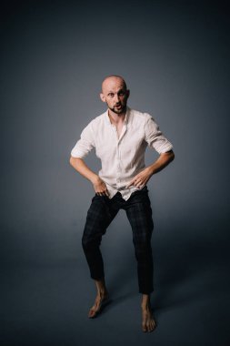 Beyaz gömlekli ve ekose pantolonlu, gri arka planda dans eden güzel sakallı, kel bir adamın stüdyo portresi.