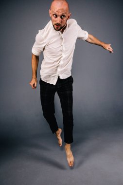 Beyaz gömlekli ve ekose pantolonlu, gri arka planda dans eden güzel sakallı, kel bir adamın stüdyo portresi.