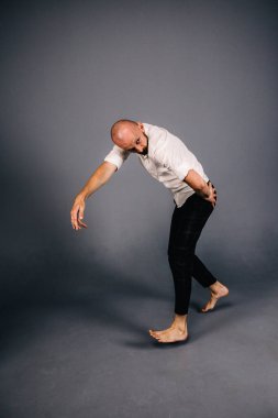 Beyaz gömlekli ve ekose pantolonlu, gri arka planda dans eden güzel sakallı, kel bir adamın stüdyo portresi.