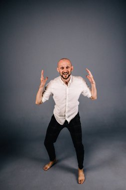 Beyaz gömlekli ve ekose pantolonlu, gri arka planda dans eden güzel sakallı, kel bir adamın stüdyo portresi.