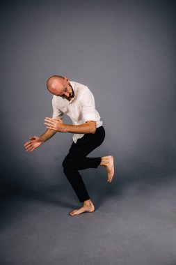 Beyaz gömlekli ve ekose pantolonlu, gri arka planda dans eden güzel sakallı, kel bir adamın stüdyo portresi.