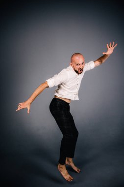 Beyaz gömlekli ve ekose pantolonlu, gri arka planda dans eden güzel sakallı, kel bir adamın stüdyo portresi.