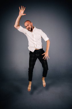 studio portrait of a bald guy with a beautiful beard barefoot in a white shirt and plaid pants dancing on a gray background