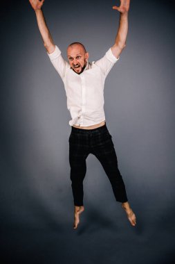 Beyaz gömlekli ve ekose pantolonlu, gri arka planda dans eden güzel sakallı, kel bir adamın stüdyo portresi.