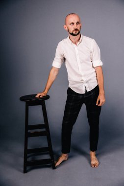 Çıplak ayaklı, kel, sakallı, beyaz gömlekli, ekose pantolonlu, gri arka planda bar taburesiyle poz veren bir adam..