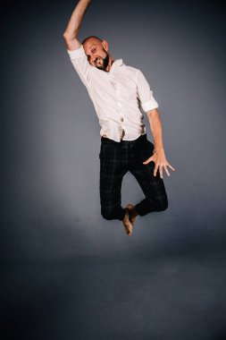 studio portrait of a bald guy with a beautiful beard barefoot in a white shirt and plaid pants dancing on a gray background