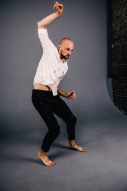 Beyaz gömlekli ve ekose pantolonlu, gri arka planda dans eden güzel sakallı, kel bir adamın stüdyo portresi.