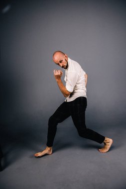 studio portrait of a bald guy with a beautiful beard barefoot in a white shirt and plaid pants dancing on a gray background