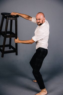 A barefoot, bald guy with a beautiful beard in a white shirt and plaid pants poses with a bar stool on a gray background.