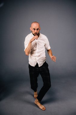 Beyaz gömlekli ve ekose pantolonlu, gri arka planda dans eden güzel sakallı, kel bir adamın stüdyo portresi.