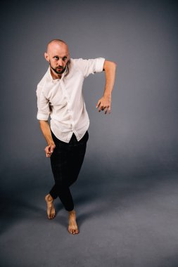 studio portrait of a bald guy with a beautiful beard barefoot in a white shirt and plaid pants dancing on a gray background