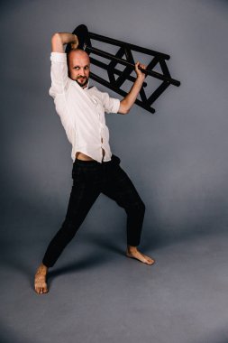 Çıplak ayaklı, kel, sakallı, beyaz gömlekli, ekose pantolonlu, gri arka planda bar taburesiyle poz veren bir adam..