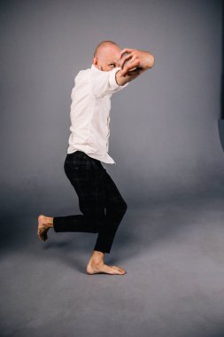 Beyaz gömlekli ve ekose pantolonlu, gri arka planda dans eden güzel sakallı, kel bir adamın stüdyo portresi.