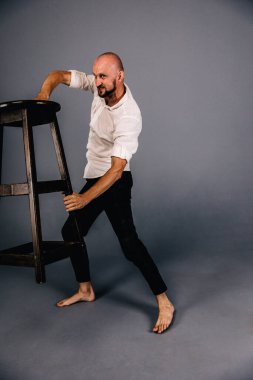 Çıplak ayaklı, kel, sakallı, beyaz gömlekli, ekose pantolonlu, gri arka planda bar taburesiyle poz veren bir adam..