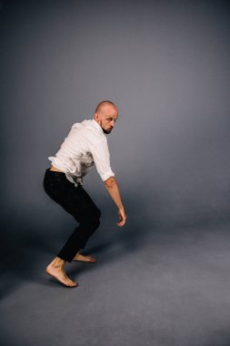 Beyaz gömlekli ve ekose pantolonlu, gri arka planda dans eden güzel sakallı, kel bir adamın stüdyo portresi.