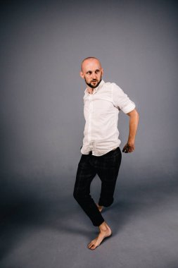 Beyaz gömlekli ve ekose pantolonlu, gri arka planda dans eden güzel sakallı, kel bir adamın stüdyo portresi.