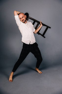 Çıplak ayaklı, kel, sakallı, beyaz gömlekli, ekose pantolonlu, gri arka planda bar taburesiyle poz veren bir adam..