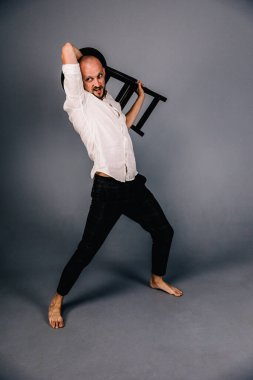 Çıplak ayaklı, kel, sakallı, beyaz gömlekli, ekose pantolonlu, gri arka planda bar taburesiyle poz veren bir adam..