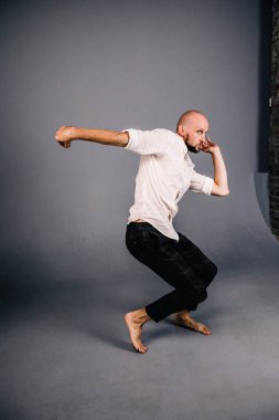 Beyaz gömlekli ve ekose pantolonlu, gri arka planda dans eden güzel sakallı, kel bir adamın stüdyo portresi.