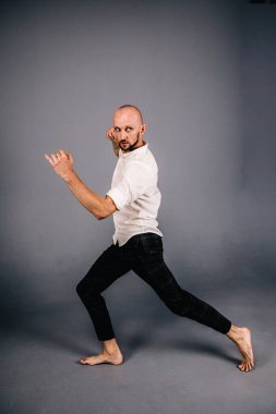 Beyaz gömlekli ve ekose pantolonlu, gri arka planda dans eden güzel sakallı, kel bir adamın stüdyo portresi.