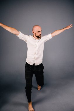 studio portrait of a bald guy with a beautiful beard barefoot in a white shirt and plaid pants dancing on a gray background