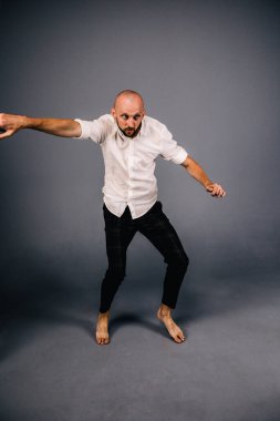 Beyaz gömlekli ve ekose pantolonlu, gri arka planda dans eden güzel sakallı, kel bir adamın stüdyo portresi.