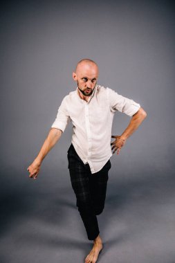 Beyaz gömlekli ve ekose pantolonlu, gri arka planda dans eden güzel sakallı, kel bir adamın stüdyo portresi.