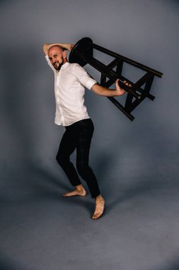 Çıplak ayaklı, kel, sakallı, beyaz gömlekli, ekose pantolonlu, gri arka planda bar taburesiyle poz veren bir adam..