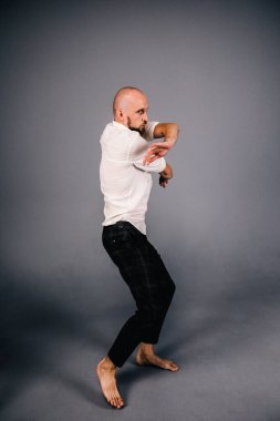 Beyaz gömlekli ve ekose pantolonlu, gri arka planda dans eden güzel sakallı, kel bir adamın stüdyo portresi.