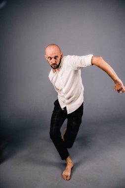 studio portrait of a bald guy with a beautiful beard barefoot in a white shirt and plaid pants dancing on a gray background
