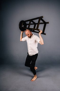 Çıplak ayaklı, kel, sakallı, beyaz gömlekli, ekose pantolonlu, gri arka planda bar taburesiyle poz veren bir adam..