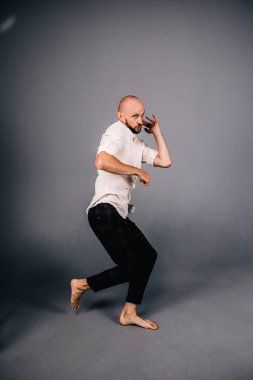 studio portrait of a bald guy with a beautiful beard barefoot in a white shirt and plaid pants dancing on a gray background