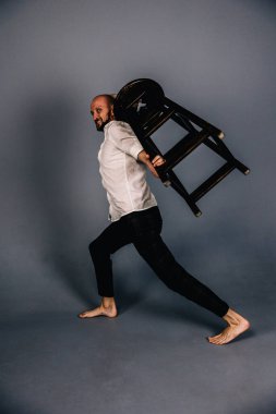 Çıplak ayaklı, kel, sakallı, beyaz gömlekli, ekose pantolonlu, gri arka planda bar taburesiyle poz veren bir adam..