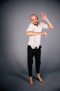 Beyaz gömlekli ve ekose pantolonlu, gri arka planda dans eden güzel sakallı, kel bir adamın stüdyo portresi.