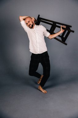 Çıplak ayaklı, kel, sakallı, beyaz gömlekli, ekose pantolonlu, gri arka planda bar taburesiyle poz veren bir adam..