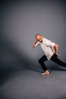 Beyaz gömlekli ve ekose pantolonlu, gri arka planda dans eden güzel sakallı, kel bir adamın stüdyo portresi.