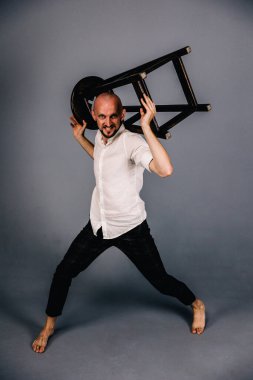 Çıplak ayaklı, kel, sakallı, beyaz gömlekli, ekose pantolonlu, gri arka planda bar taburesiyle poz veren bir adam..