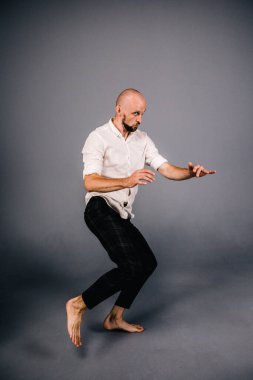 Beyaz gömlekli ve ekose pantolonlu, gri arka planda dans eden güzel sakallı, kel bir adamın stüdyo portresi.
