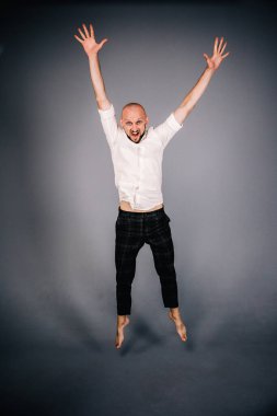 Beyaz gömlekli ve ekose pantolonlu, gri arka planda dans eden güzel sakallı, kel bir adamın stüdyo portresi.