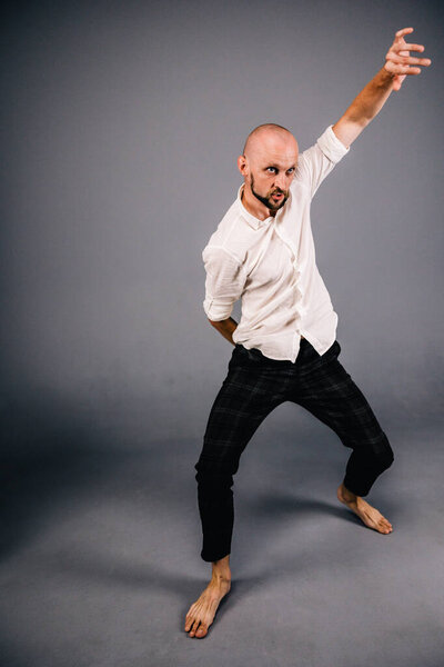 studio portrait of a bald guy with a beautiful beard barefoot in a white shirt and plaid pants dancing on a gray background
