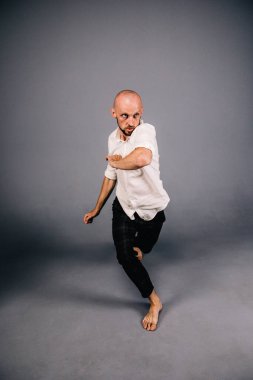Beyaz gömlekli ve ekose pantolonlu, gri arka planda dans eden güzel sakallı, kel bir adamın stüdyo portresi.