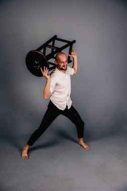 Çıplak ayaklı, kel, sakallı, beyaz gömlekli, ekose pantolonlu, gri arka planda bar taburesiyle poz veren bir adam..